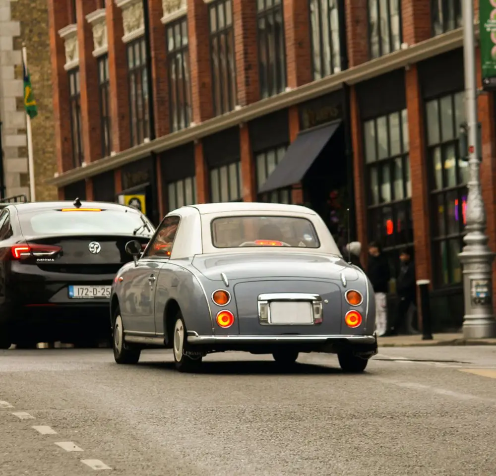 A small, vintage blue car with a white roof—perfect for Nissan Figaro Insurance—drives on a city street beside a black 4x4, with brick buildings and shops in the background.