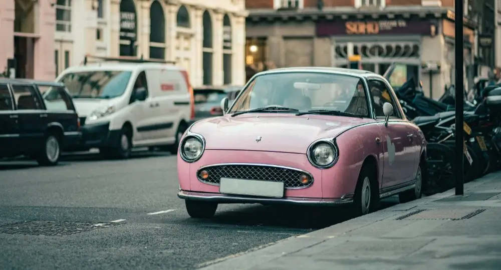 A vintage pink Nissan Figaro is parked on a city street next to the pavement, with other vehicles and buildings visible in the background—a perfect scene for anyone seeking Nissan Figaro insurance.