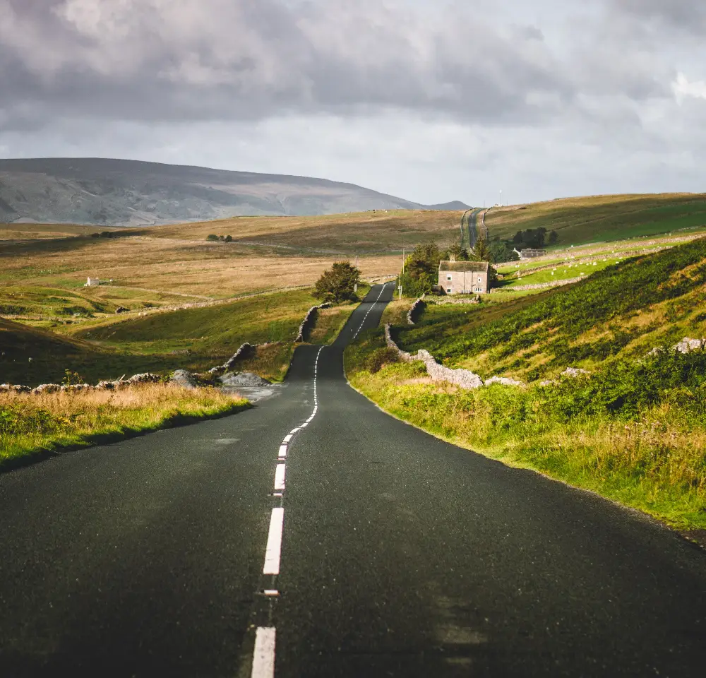 A long, empty country road stretches into rolling green hills under a cloudy sky, passing a stone house surrounded by fields and low stone walls—an ideal scene for peace of mind with Westfield car insurance on your journey.