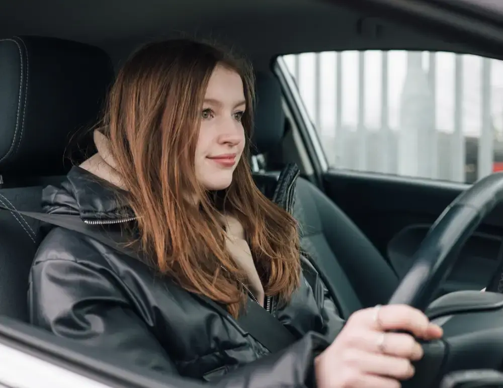 A young woman with long brown hair, wearing a black jacket, sits in the driver's seat of a car—possibly considering Mitsubishi insurance—as she looks ahead whilst holding the steering wheel.