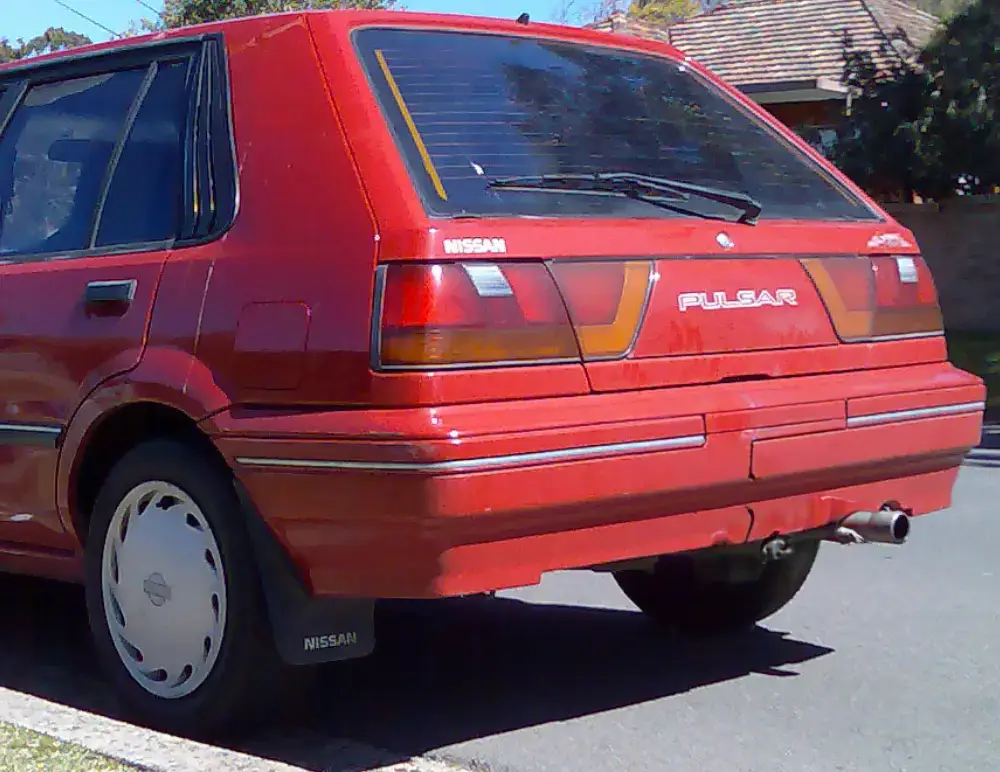 A red Nissan Pulsar hatchback is parked on the street, viewed from the rear left side. The car has a boxy shape, tinted rear window, visible Nissan and Pulsar badges—make sure your Car Insurance covers every journey.