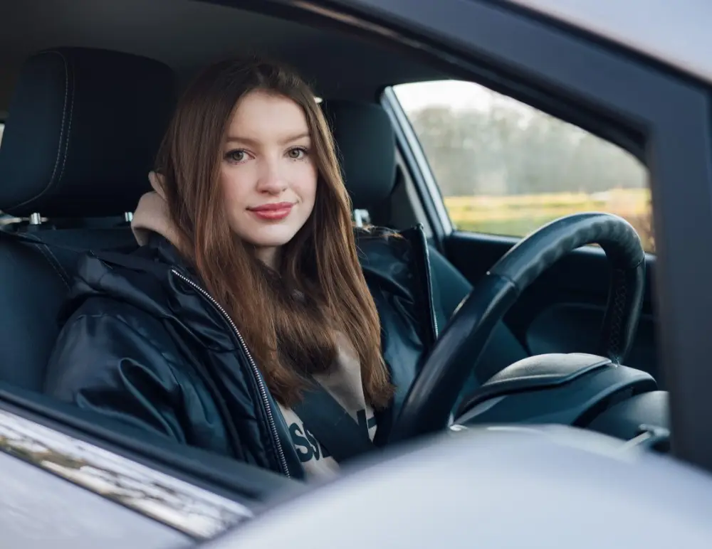 A young woman with long brown hair sits in the driver’s seat of a Nissan Pulsar, looking at the camera with a slight smile. She is wearing a black jacket and beige hoodie. The photo is taken from outside the vehicle—perfect for a Nissan Pulsar Insurance advert.