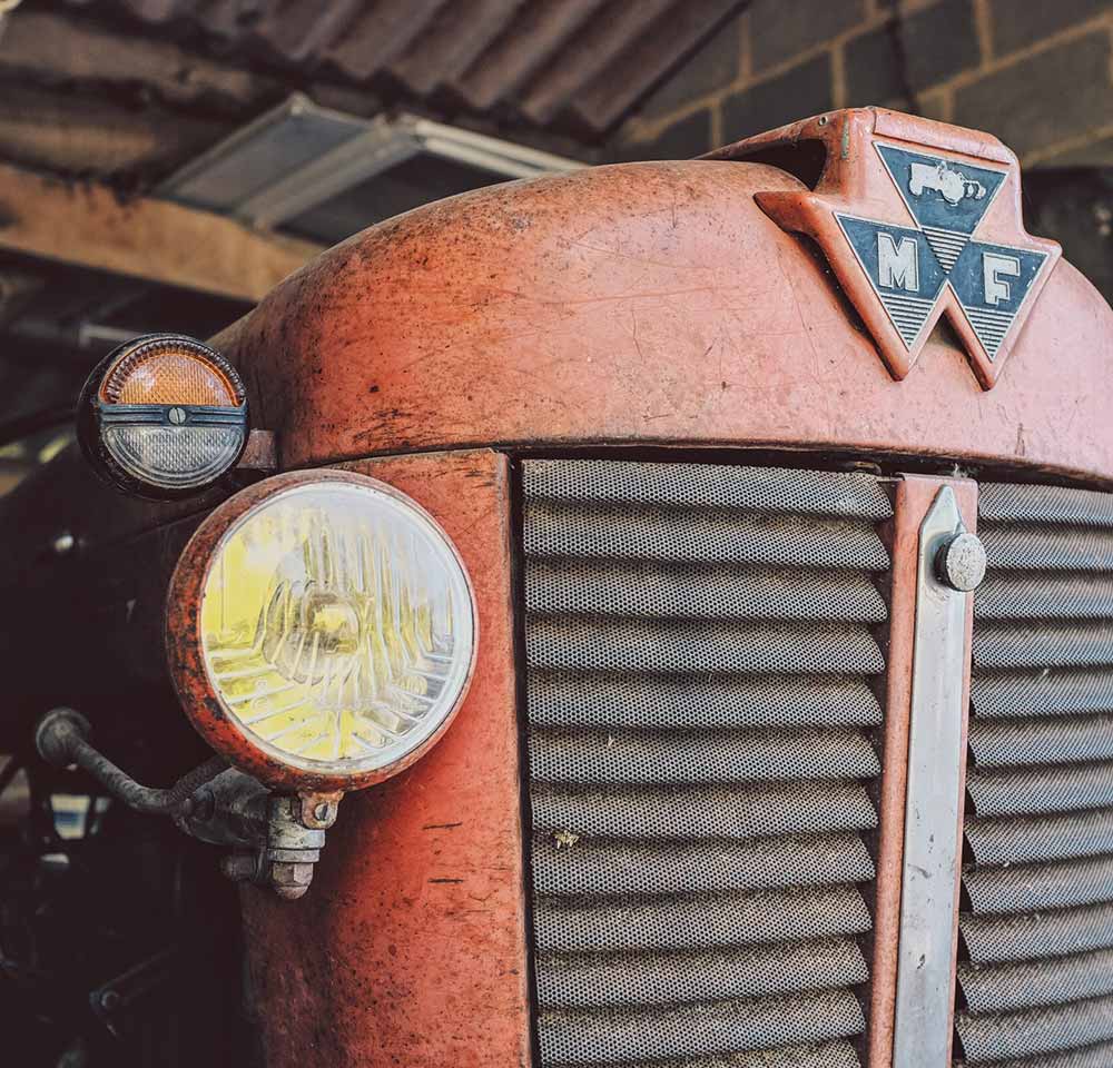 Close-up of the front of a vintage red Massey Ferguson tractor, showcasing its headlamp, grille, and MF badge on the bonnet in a rustic setting—perfect for those interested in Vintage Tractor Insurance.
