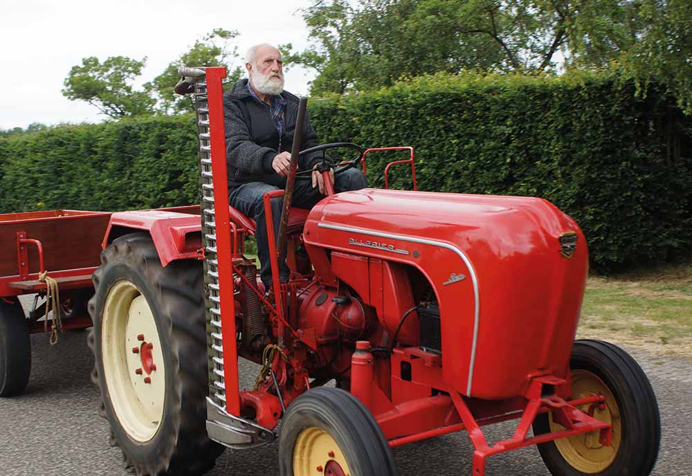 An older man with a white beard drives a vintage red tractor, covered by Classic Tractor Insurance, pulling a trailer on a tarmacked road, with green hedges and trees in the background.