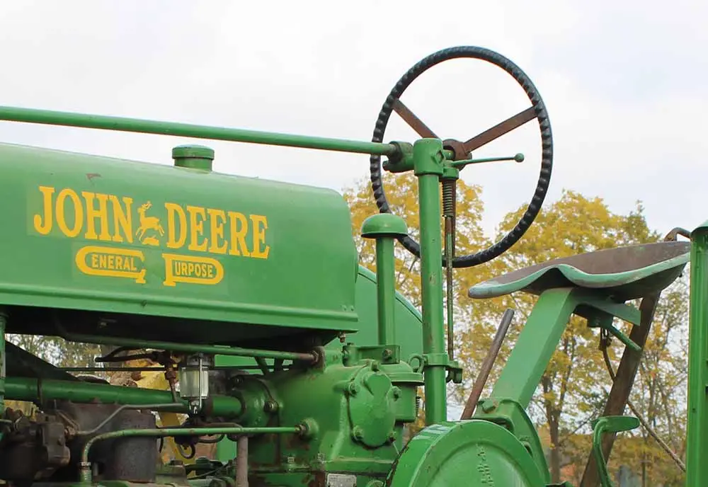 Close-up of a vintage green John Deere tractor, showing the steering wheel, metal seat, and iconic yellow brand name—an ideal candidate for Classic Tractor Insurance. Trees with autumn foliage appear in the blurry background.