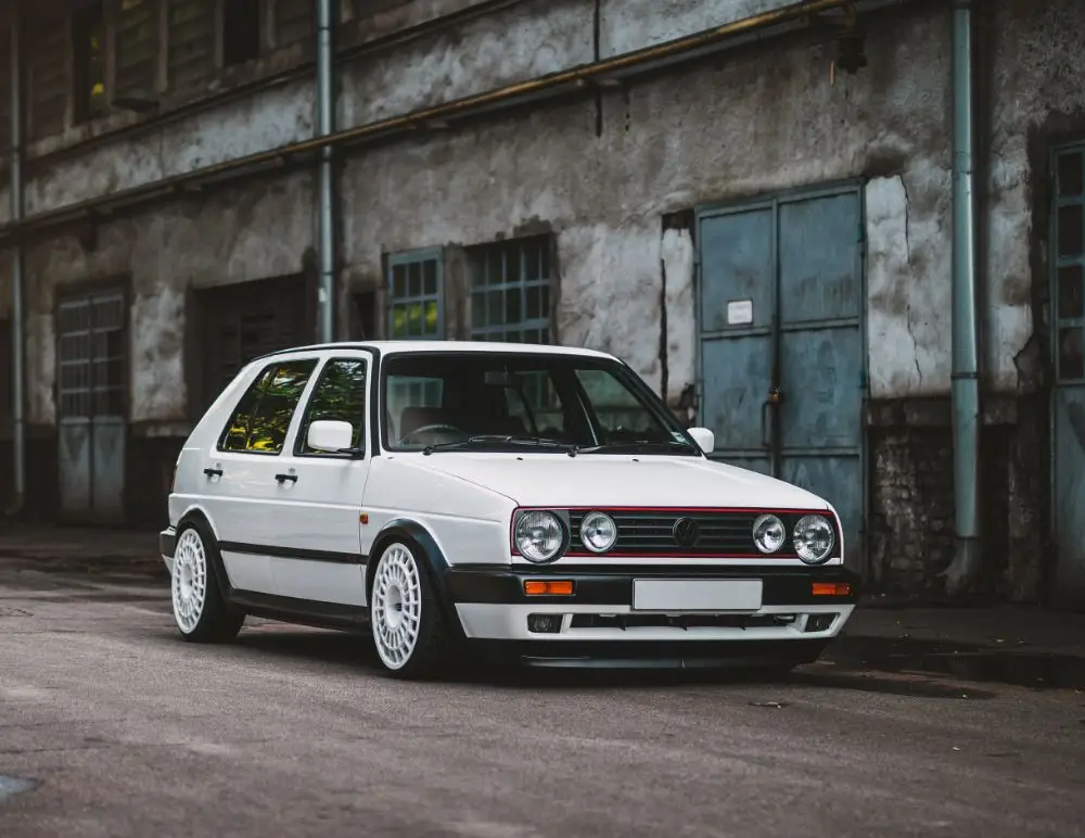 A white Volkswagen Golf Mk2 hatchback with custom wheels is parked on a street beside an old, weathered industrial building—an ideal scene for anyone searching for the perfect VW Golf Insurance option.