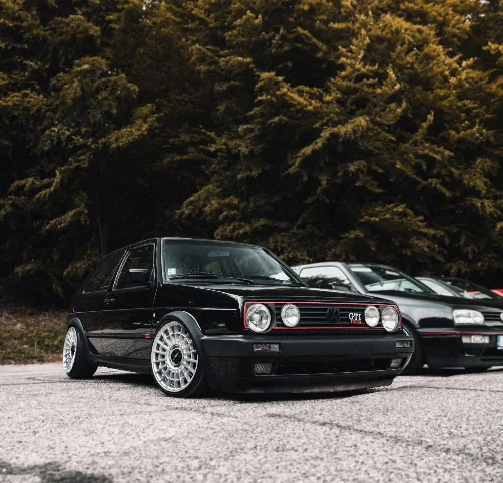 A black Volkswagen Golf GTI with custom wheels is parked on a paved surface, surrounded by trees and another car in the background—an ideal scene to consider VW Golf Insurance for your prized motor at a car meet or outdoor gathering.