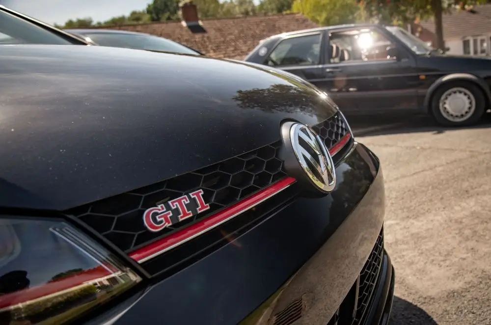 Close-up of the front grille of a black Volkswagen GTI with red detailing and GTI badge, perfect for those considering VW Golf Insurance, parked next to another black car in a sunny outdoor setting with trees and houses in the background.