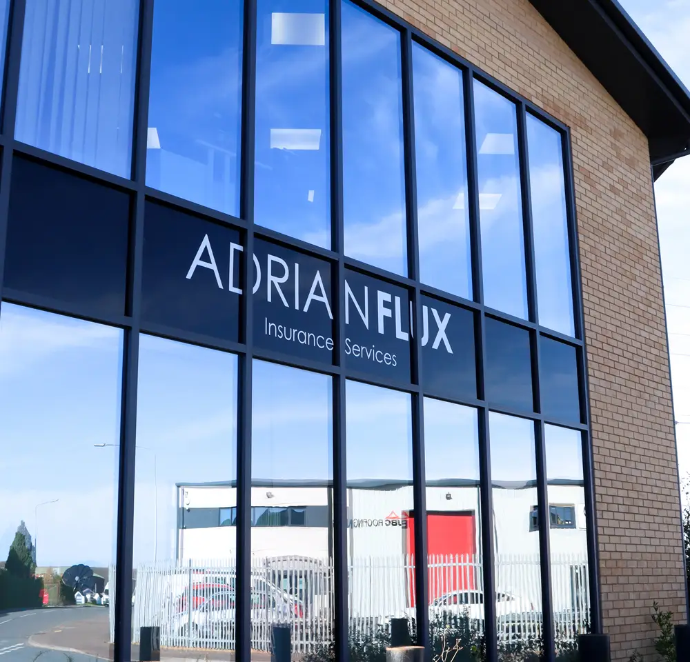 The exterior of a modern building with large reflective windows displays the sign Adrian Flux Insurance Services, specialists in Reliant Robin insurance. The reflection shows nearby buildings and a cloudy blue sky.