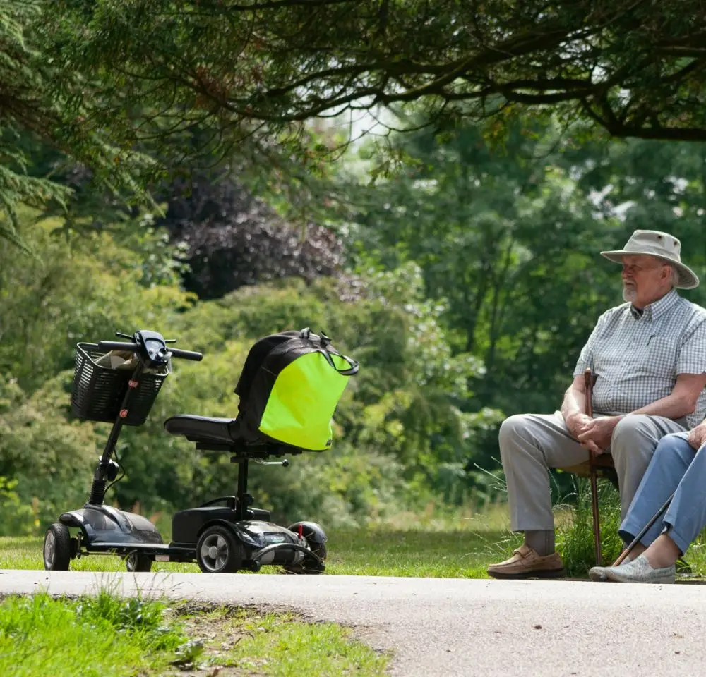 Two elderly people sit on a bench in a park, shaded by trees. In front of them is a mobility scooter, covered by Mobility Scooter Insurance, with a bright yellow bag attached. Lush greenery surrounds the peaceful scene.