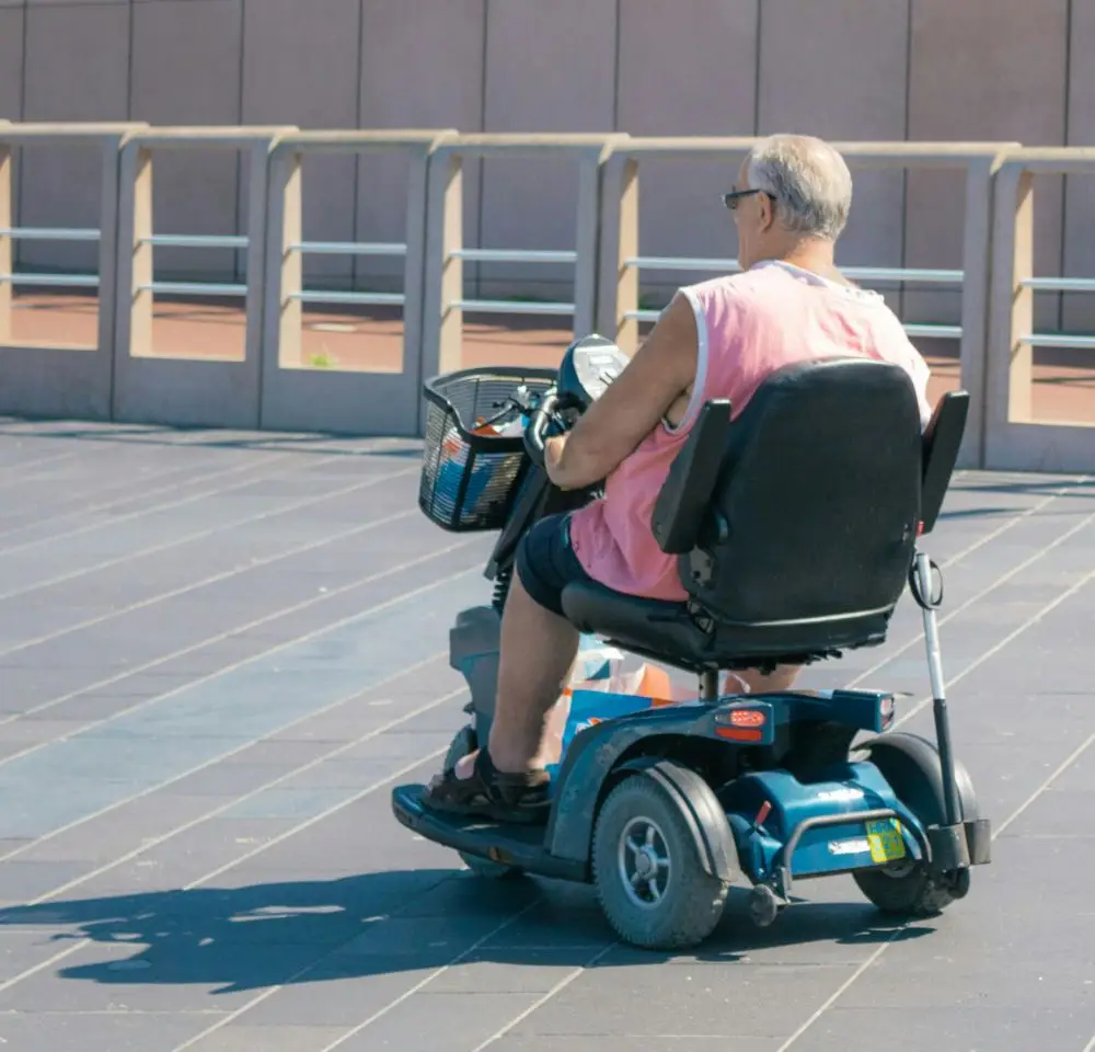 An older man in a sleeveless pink shirt and sunglasses rides a blue mobility scooter on a paved area outdoors, with a modern railing and building in the background—Mobility Scooter Insurance can help keep such journeys worry-free.