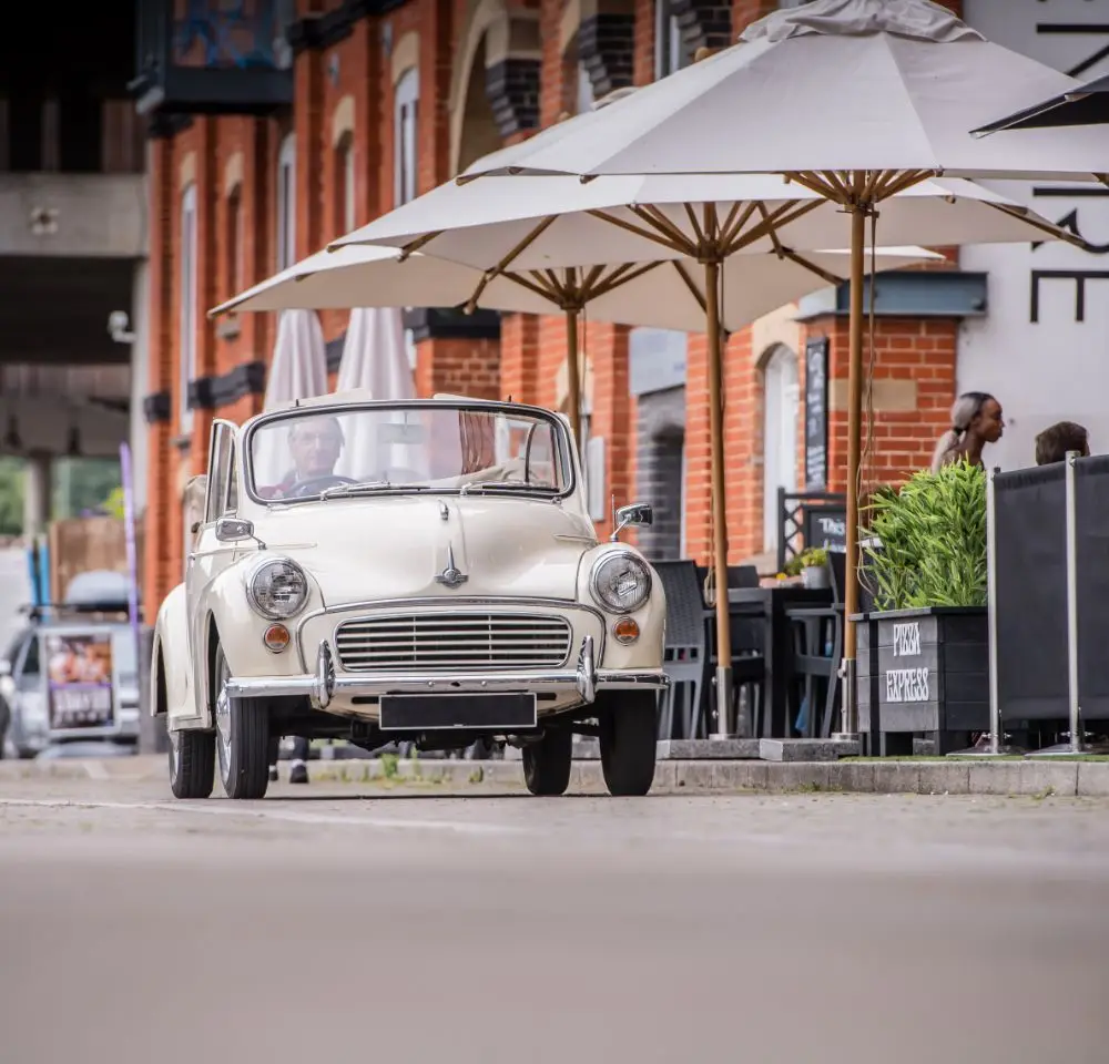 A classic cream-coloured convertible car, ideal for Morris Minor Insurance, drives past a brick building with outdoor seating and large white umbrellas; people sit and relax at tables along the pavement.