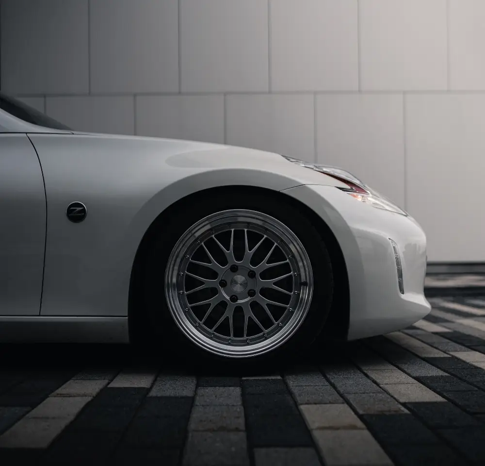 Close-up side view of a white Nissan 350Z sports car parked on patterned stone pavement, featuring a shiny alloy wheel and part of the front bumper against a plain wall background.