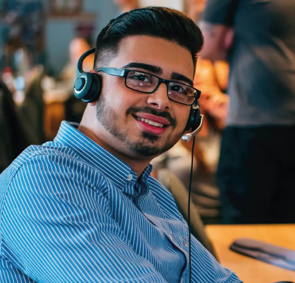 A young man wearing glasses and a headset smiles whilst sitting at a desk in an office environment. He is dressed in a blue striped shirt. The background shows other people working.