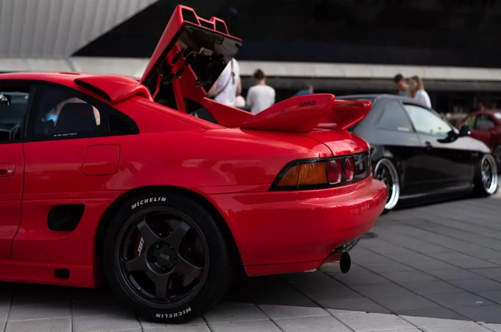 A bright red sports car with its rear engine cover open is parked on a tiled surface, possibly showcasing Toyota MR2 insurance at a car meet or exhibition, with another car and several people visible in the background.