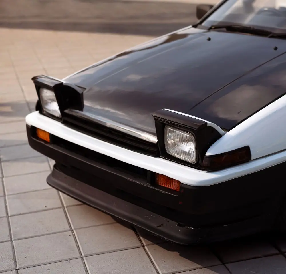 Close-up of the front of a classic Toyota MR2 with pop-up headlamps raised, black bonnet, and white bumper, parked on light-coloured tiled pavement—ideal for those considering Toyota MR2 insurance.