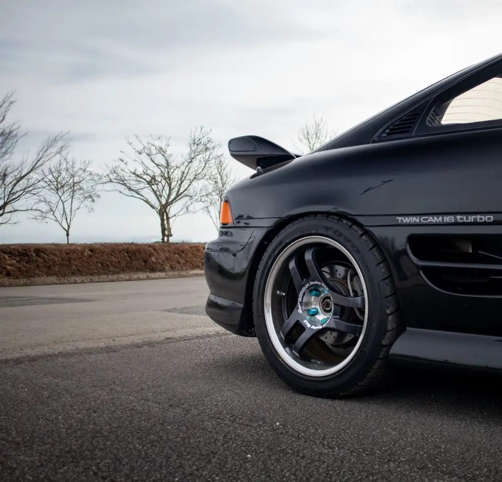 Close-up of the rear side of a black sports car, possibly covered by Toyota MR2 insurance, parked on a road with sporty wheels and a spoiler visible. Bare trees and a cloudy sky are in the background.