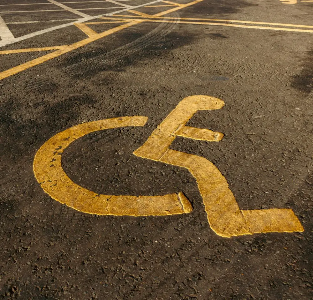 Yellow wheelchair accessible symbol painted on asphalt marks a reserved parking space for people with disabilities. Faded parallel yellow lines are visible in the background, highlighting access for wheelchair accessible vehicles.