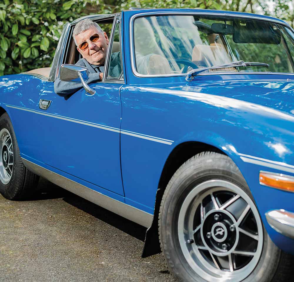 An older man wearing sunglasses smiles whilst sitting in the driver’s seat of a classic blue convertible car, with Triumph insurance ensuring his peace of mind as he enjoys the day outdoors.