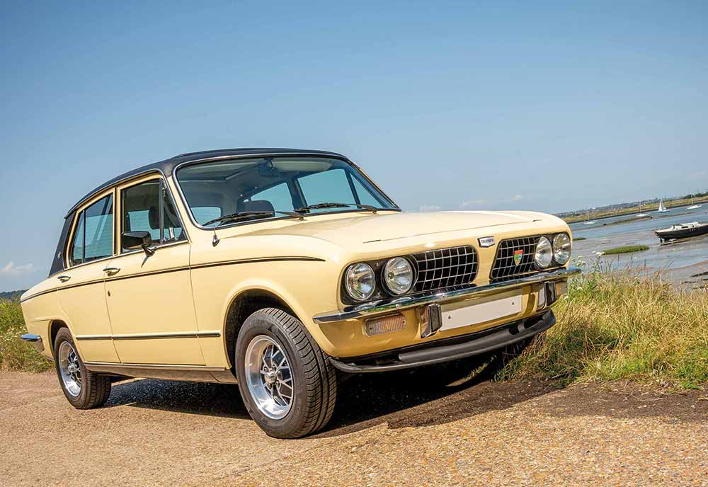 A vintage cream-coloured saloon with chrome trim and classic round headlamps is parked on a rural road beside grassy terrain and water under a clear blue sky, perfectly capturing the spirit of adventure—and peace of mind with Triumph insurance.