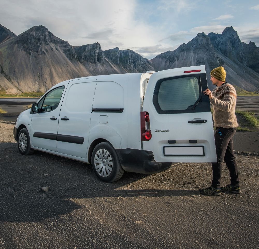 Person in a yellow beanie and knitted sweater opening the rear door of a white Citroën Berlingo van parked on a gravel surface in a mountainous landscape, with dramatic peaks and a partly cloudy sky in the background.
