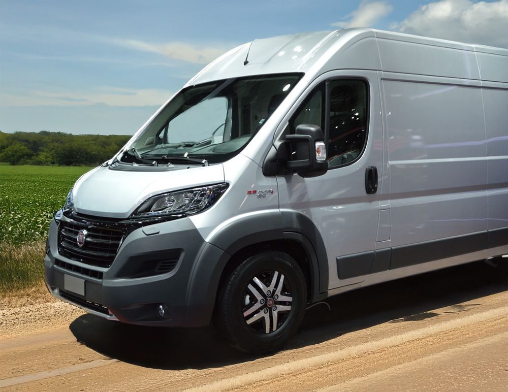 Silver Fiat van parked on a dirt road beside a green field under a blue sky with scattered clouds.