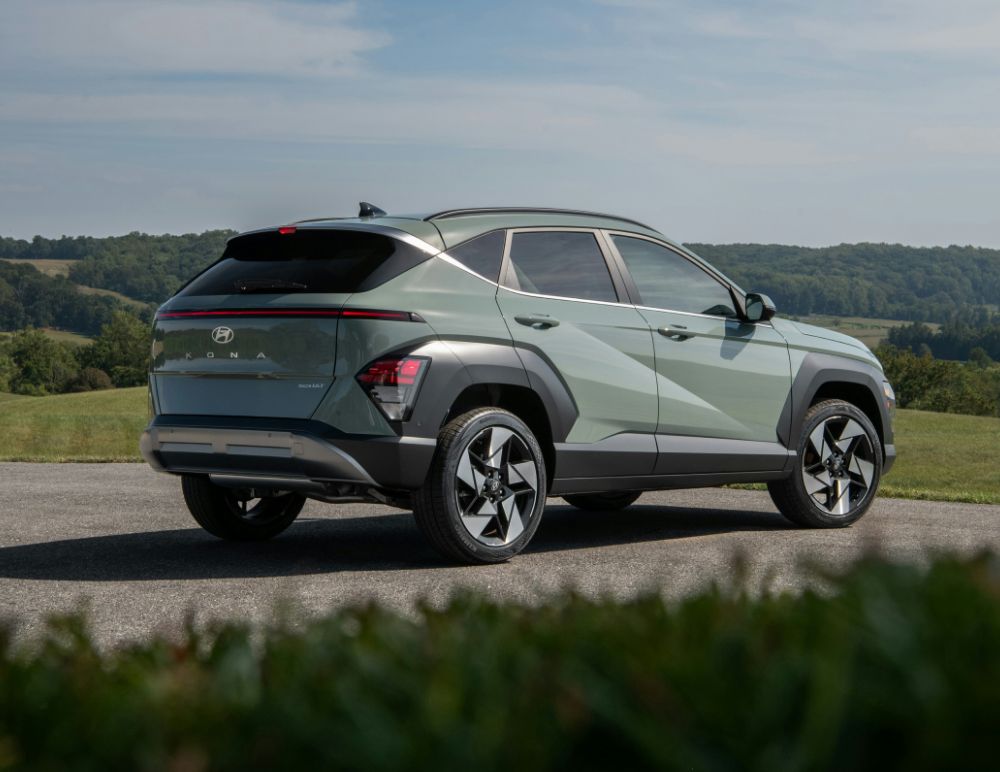 A Hyundai Kona compact SUV is parked on a paved surface with rolling green hills in the background. The vehicle is shown from a rear three-quarter angle, emphasizing its angular body lines, sleek rear light bar, and modern alloy wheels.